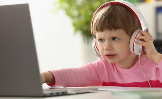 Little girl child in headphones in front of laptop. Children emotions of discontent and focusing attention concept