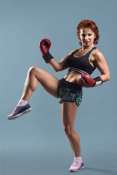 Mature athletic redhead woman in sportswear t-shirt and shorts doing fitness exercises in boxing gloves, gray studio background
