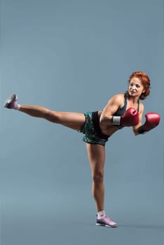 Mature athletic redhead woman in sportswear t-shirt and shorts doing fitness exercises in boxing gloves, gray studio background