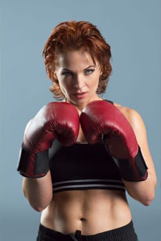 Studio portrait of sporty mature woman in boxing gloves on gray background, athletic red-haired serious female looking at camera