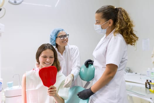 Teenager girl in dental chair with mirror looking at her teeth, smiling doctor and assistant. Medicine, dentistry and healthcare concept