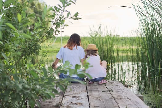 Children resting near the water on sunny summer day, two girls looking at lake sitting on bridge, relaxing, copy space, back view