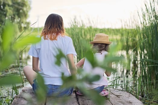 Children resting near the water on sunny summer day, two girls looking at lake sitting on bridge, relaxing, back view