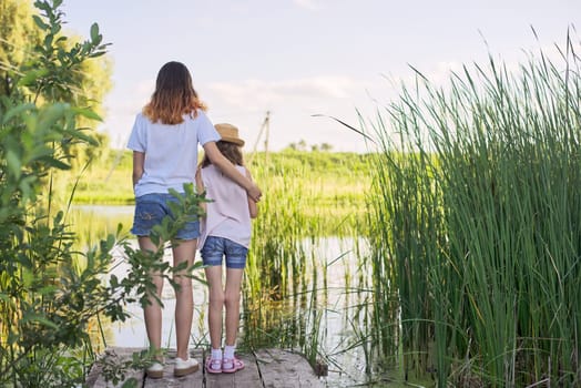 Children resting near the water on sunny summer day, two girls looking at lake on bridge, relaxing, copy space, back view