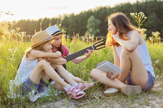 Children sitting in nature with classical guitar, three girls learning playing the guitar and singing, enjoying music and summer day