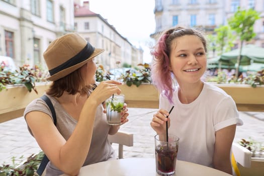 Two women mother and teenage daughter sitting in city outdoor cafe talking, drinking summer ice cold drinks, mojito, cocktail