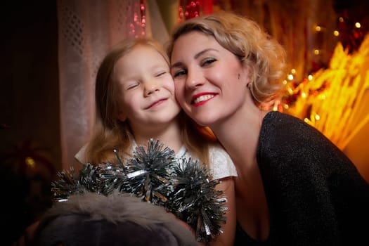 Cute mother and daughter in a room with Christmas garlands. The tradition of decorating the house and dressing up for the holidays. Gifts for family. Happy childhood and motherhood