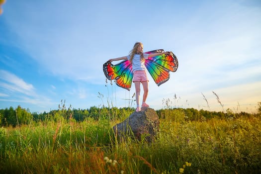 Pretty blonde girl with bright butterfly wings having fun in meadow on natural landscape with grass and flowers on sunny summer day. Portrait of teenage child in spring season outdoors on field