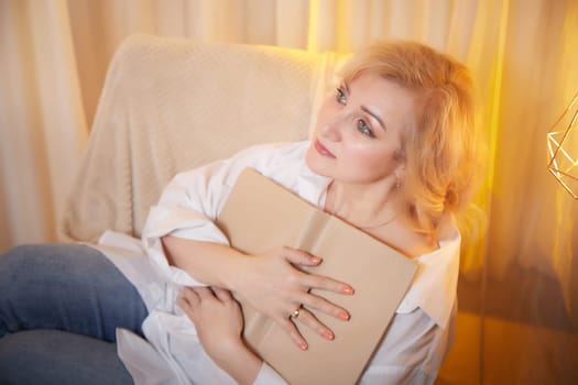 Adult mature woman of 40 or 60 years old with book in casual dress white shirt and blue jeans in calm cozy evening atmosphere room. Interior with transparent curtains and soft warm lamp