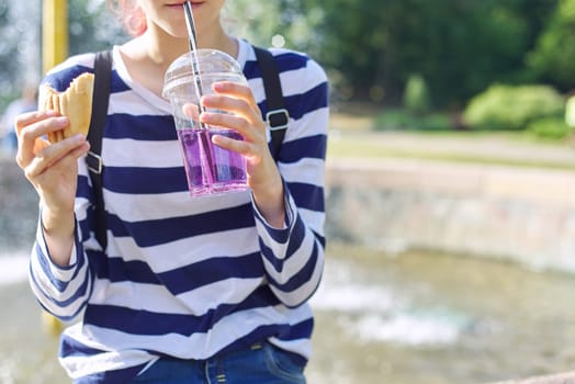 Street food, teenager girl eating sandwich and drinking drink from glass with straw.