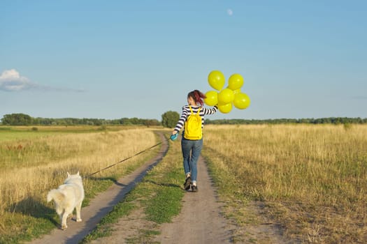 Dynamic outdoor portrait of running girl with white dog and yellow balloons on country road, beautiful landscape with blue cloudy sky and yellow grass in the meadow. Back view