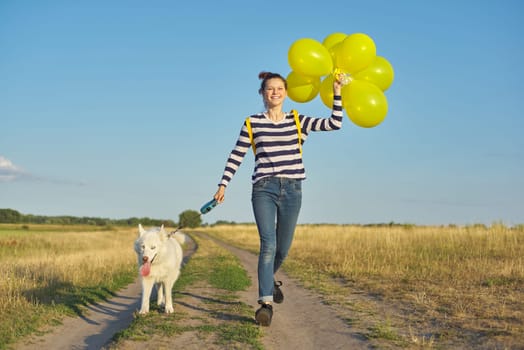 Dynamic outdoor portrait of running girl with white dog and yellow balloons on country road, beautiful landscape with blue cloudy sky and yellow grass in the meadow