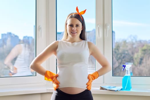 House cleaning, young beautiful pregnant woman in gloves with rag and detergent posing, looking at camera, near clean washed window in room