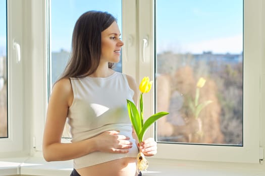 Spring, beauty, health, portrait of young pregnant woman with yellow tulip at home near window. Beautiful female with healthy skin and hair, natural makeup, copy space