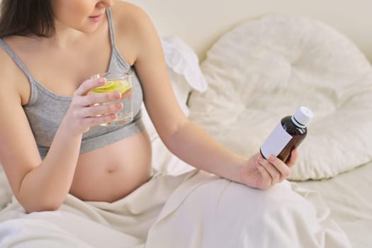 Pregnant woman reading label on bottle with medicine, with vitamins. Female sitting at home in bed with glass of water medicine. Pregnancy, health, pharmaceuticals, care and people.
