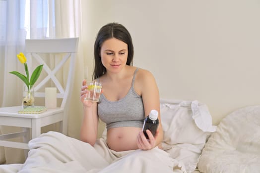 Pregnant woman reading label on bottle with medicine, with vitamins. Female sitting at home in bed with glass of water medicine. Pregnancy, health, pharmaceuticals, care and people.