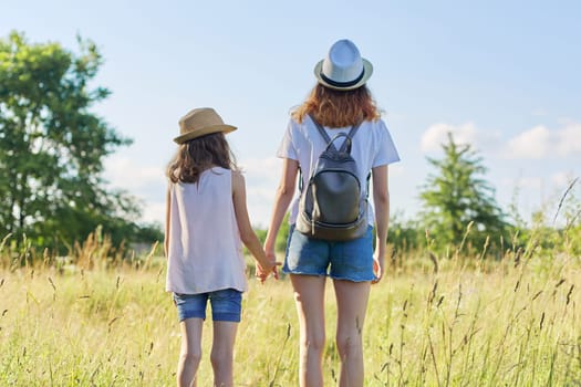 Summer vacation, children two girls sisters walking together holding hands, blue sky meadow background. Kids in hats with backpacks, back view