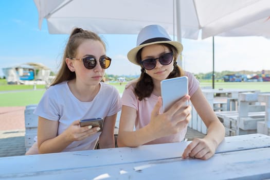 Two teenage girls with smartphones sitting and talking in summer outdoor cafe. Youth, teens, friendship, communication, people concept