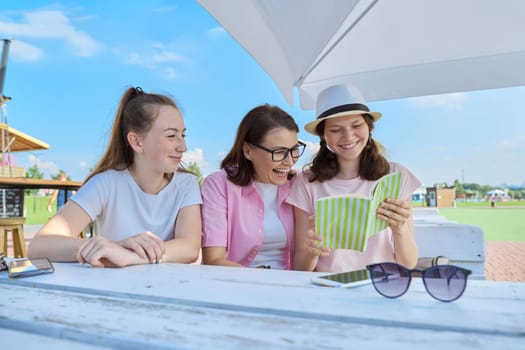 Mom and daughters teenagers have fun, look and read funny book. Communication of parent and children of adolescents. Sit in street cafe, sunny summer day