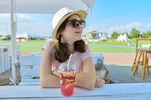 Summer fashion portrait of teenage girl in sunglasses hat sitting in outdoor cafe with refreshing berry cocktail. Vacation, rest, youth, summertime