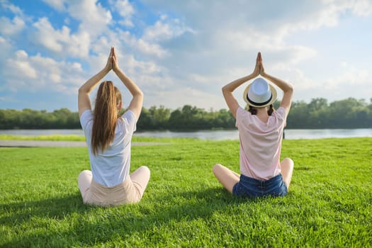 Two girls sitting in lotus position on green grass, back view, meditation on beautiful landscape, background blue sky in clouds, copy space