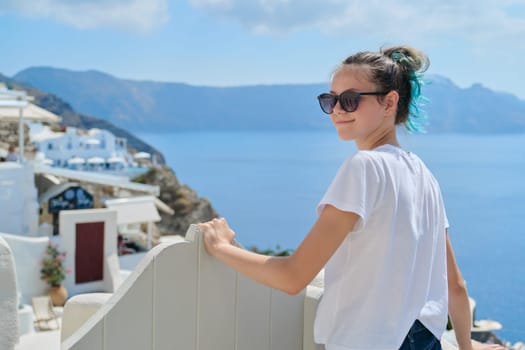 Teenager girl resting on Greek island Santorini, female looking at camera, background white architecture of village Oia, sea, sky in clouds, copy space