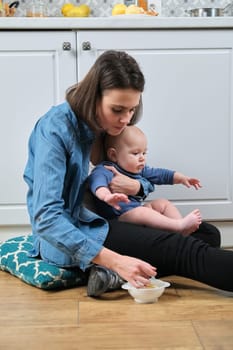 Young mother holding baby in arms, feeds toddler son with spoon. First meal, vegetable fruit puree. Woman with baby at home in kitchen