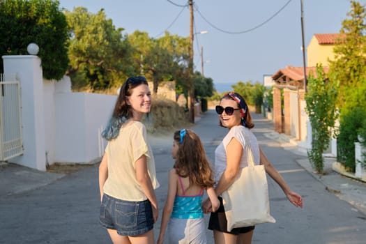 Mother and two daughters teenager and youngest walking together holding hands. Friendly family, happy parent and children, summer resort landscape background, back view