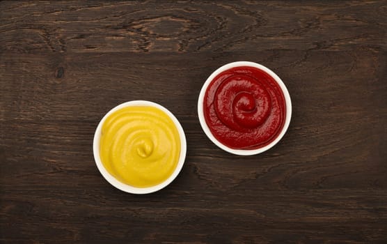 Close up two white ceramic bowls of red ketchup tomato sauce and yellow mustard on brown table surface background with copy space, top view, directly above