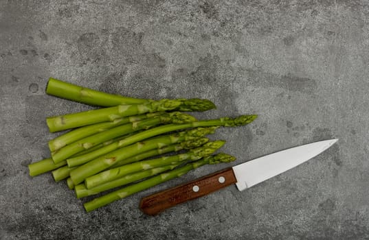 Close up bunch of fresh green asparagus and knife on cutting board or grey stone table surface, elevated top view, directly above