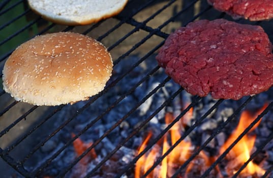 Close up raw beef meat burgers and sesame buns for hamburger cooked on fire barbecue grill with smoke and flame, high angle view