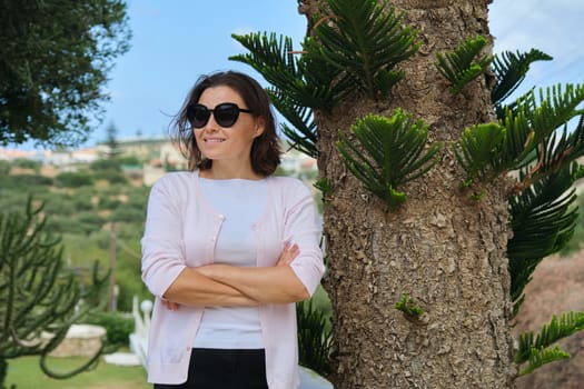 Confident beautiful mature businesswoman owner of tourist business, hotel posing with folded arms. Background resort landscape, tropical plants, copy space