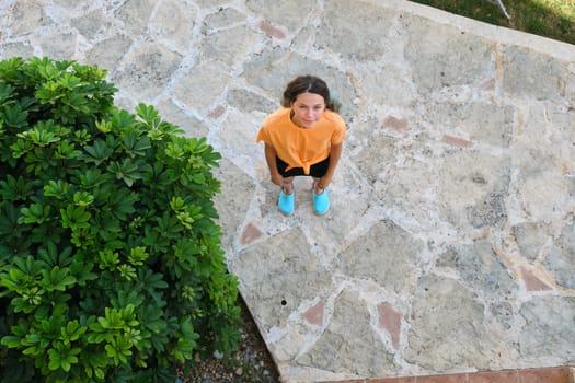 Smiling teenager girl looking up in camera, top view. Sunny summer day, female standing on gray stone sidewalk, background, copy space