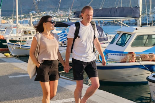 Middle-aged man and woman walking together holding hands. Love, romance, communication mature people. Background summer seascape, moored yachts in bay