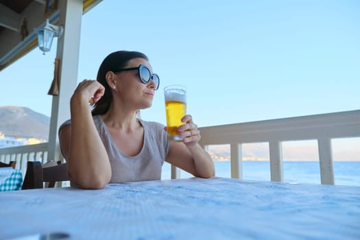 Mature beautiful woman resting sitting in sea cafe drinking cold glass of beer. Summer vacation, female enjoys sea sunset landscapes and delicious drink, copy space