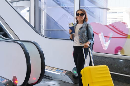 Female passenger in inside airport terminal with luggage, suitcase and backpack, traveling by air, woman near escalator holding documents and tickets, copy space