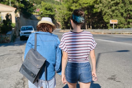 Two women mother and daughter teenager walking on sunny summer day on mountain road. Tourism, excursions, travel, family vacations, back view