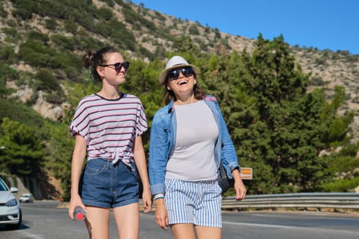 Two women mother and daughter teenager walking on sunny summer day on mountain road. Tourism, excursions, travel, family vacations