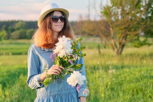 Portrait of young smiling beautiful girl in sunglasses hat with bouquet of wildflowers, sunset in meadow, copy space, rural scenery, golden hour
