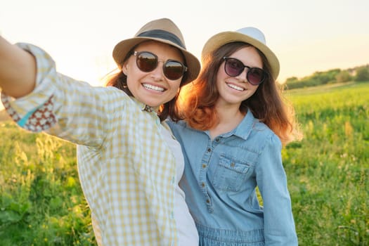 Happy smiling mother and teen daughter taking selfie photo. Females walking in meadow on sunny summer day. Parent child relationship