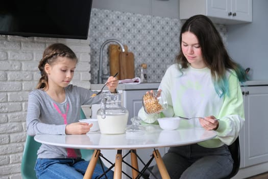 Girls having breakfast sitting at table in home kitchen, teenage sisters and 9, 10 year old child eating cornflakes with milk together. Family, food, communication, health concept.