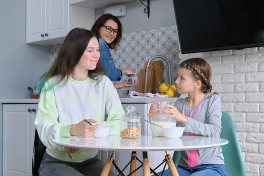 Girls having breakfast sitting at table in home kitchen, teenage sisters and 9, 10 year old child eating cornflakes with milk together. Family, food, communication, health concept.