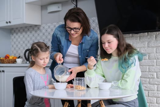 Happy mother caring for daughters in kitchen. Eating girls sitting at table, morning breakfast, milk in jug and sweet corn flakes. Family, eating, communication, health concept