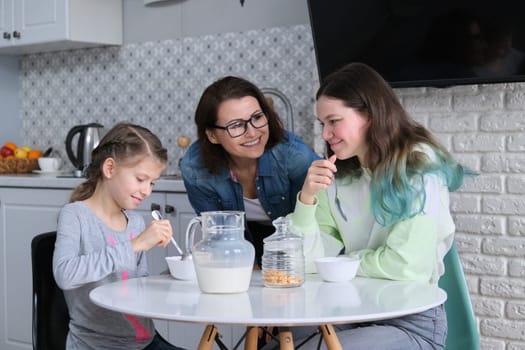 Happy mother caring for daughters in kitchen. Eating girls sitting at table, morning breakfast, milk in jug and sweet corn flakes. Family, eating, communication, health concept