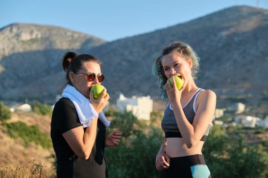 Family mother and daughter teenager in sportswear eating apples after exercise, jogging in nature, sunny day in mountains. Active healthy lifestyle and food, natural vitamins, diet products