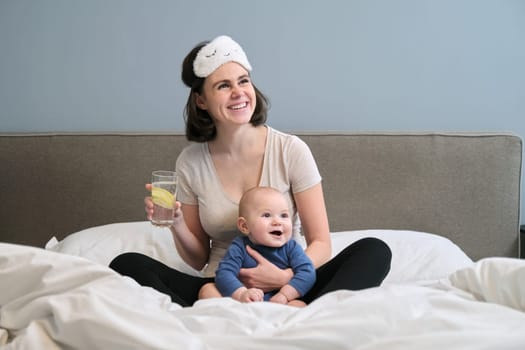 Young woman with baby toddler in arms drinking clear water with slice of lemon, sitting at home in bed. Healthy food drink for mother and baby