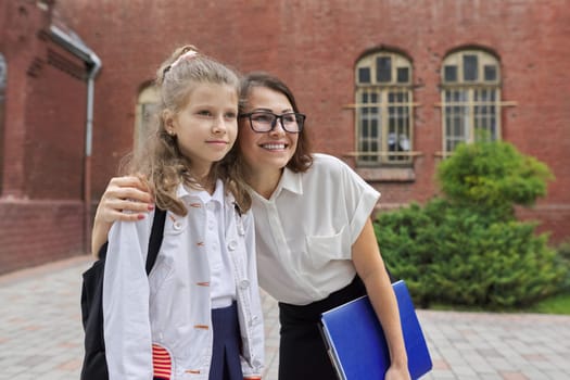 Outdoor portrait of teacher woman and little student girl together. Teacher hugging child near school building. Back to school, start of classes