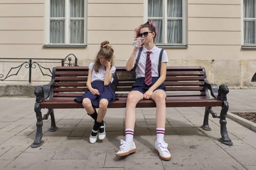 Tired schoolchildren sitting on bench. Two girls sisters teenager and elementary school student resting after class at school. Fatigue, stress, problems in school life