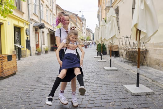 Two girls sisters having fun on the way to school. Teenager girl is circling younger, children are laughing, sunny summer day in city