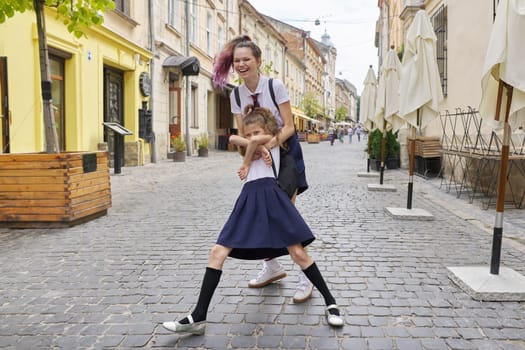 Two girls sisters having fun on the way to school. Teenager girl is circling younger, children are laughing, sunny summer day in city